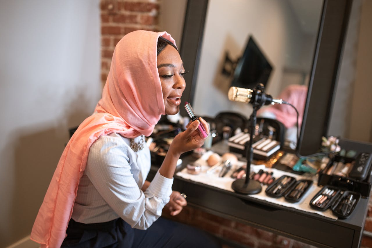 about-us Muslim woman in pink hijab applying makeup while recording a beauty vlog indoors.